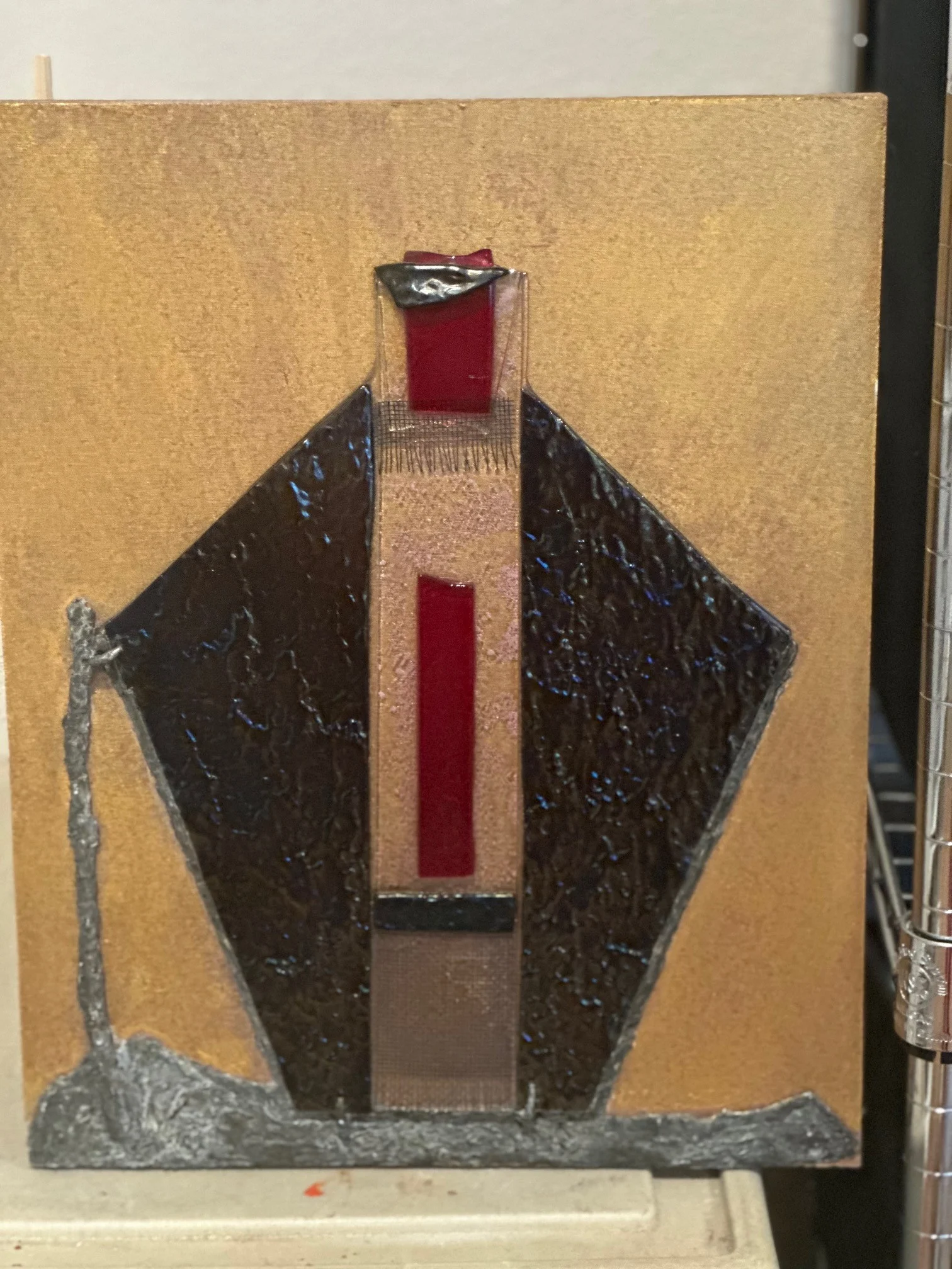 Fused glass abstract priest figure in dark glass with red accents on a gold board